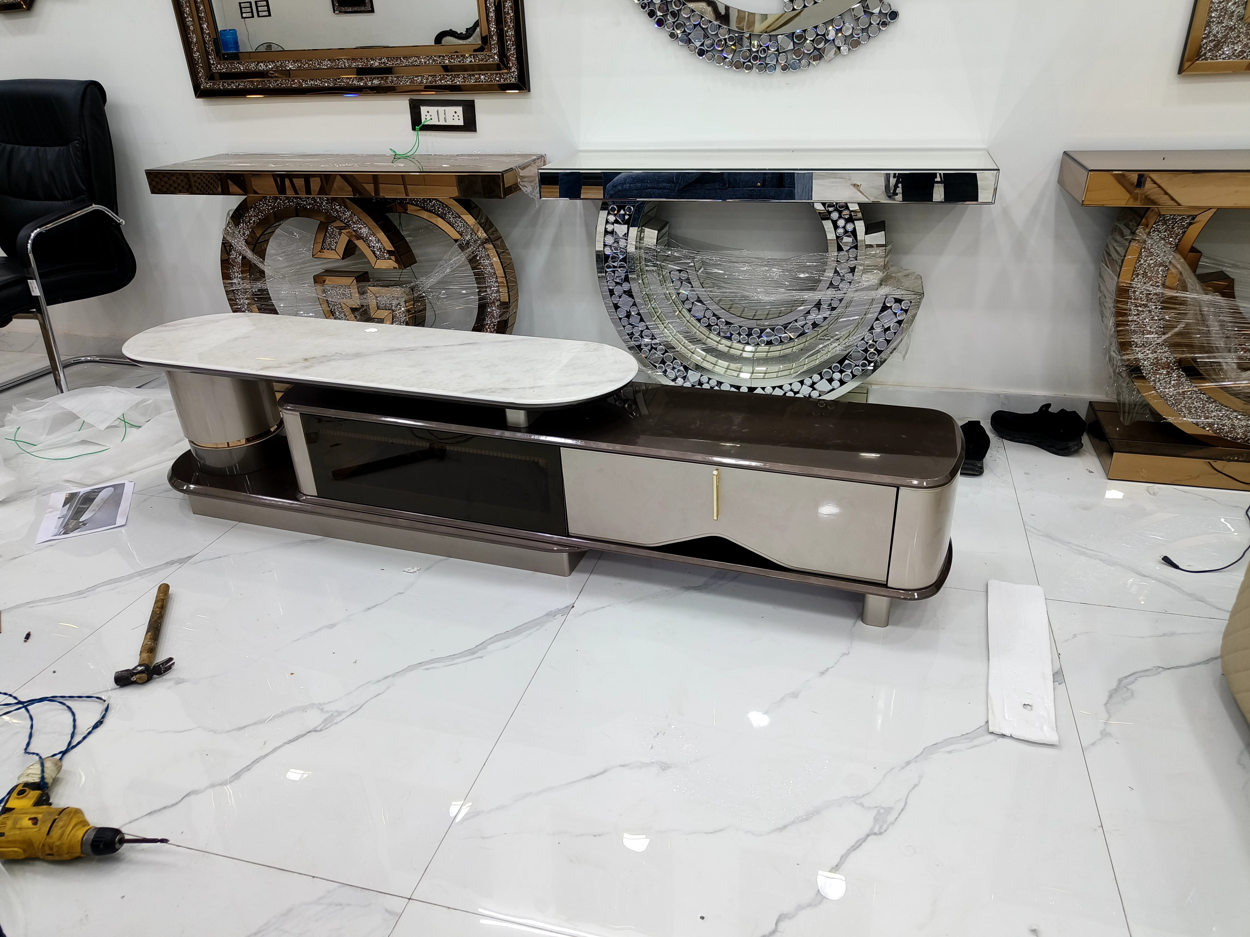 A TV cabinet and mirrored console tables with a drill, hammer, and packing materials on the tiled floor.